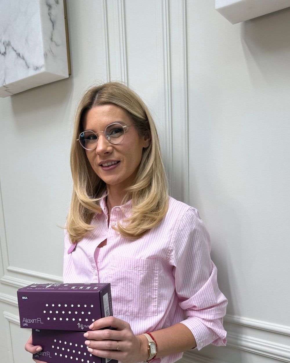 Portrait: Dr Rada at the clinic, blonde hair and glasses, pink striped shirt, holding two boxes of Aliaxin filler